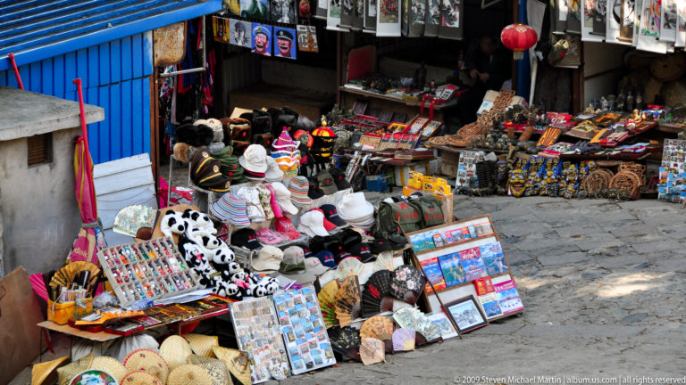 Densely stocked outdoor souvenir stall at a Great Wall visitor area, with panda plush toys, hats, postcards, and other tourist merchandise on display.