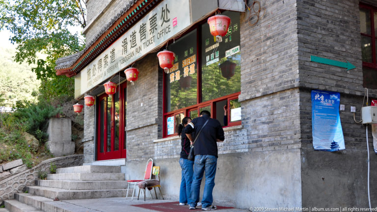 Visitors at the Mutianyu Great Wall toboggan ticket office in a gray-brick building with red lanterns and bilingual signage.