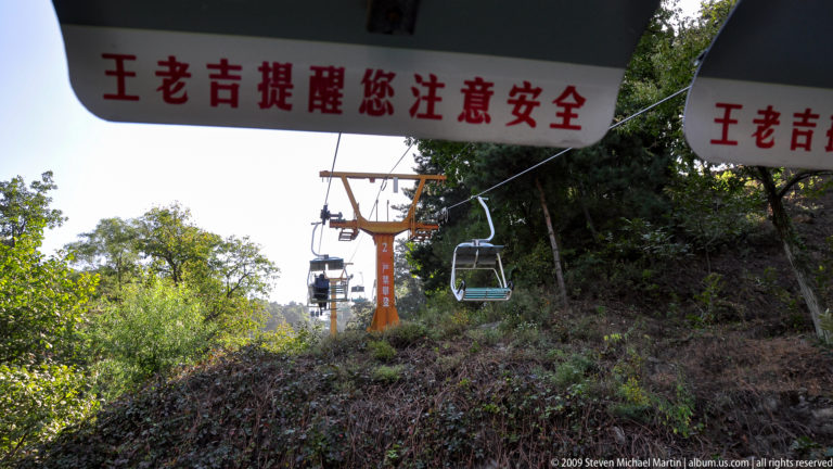 Open chairlift passing an orange support tower and Chinese safety warning sign above a wooded hillside at Mutianyu.