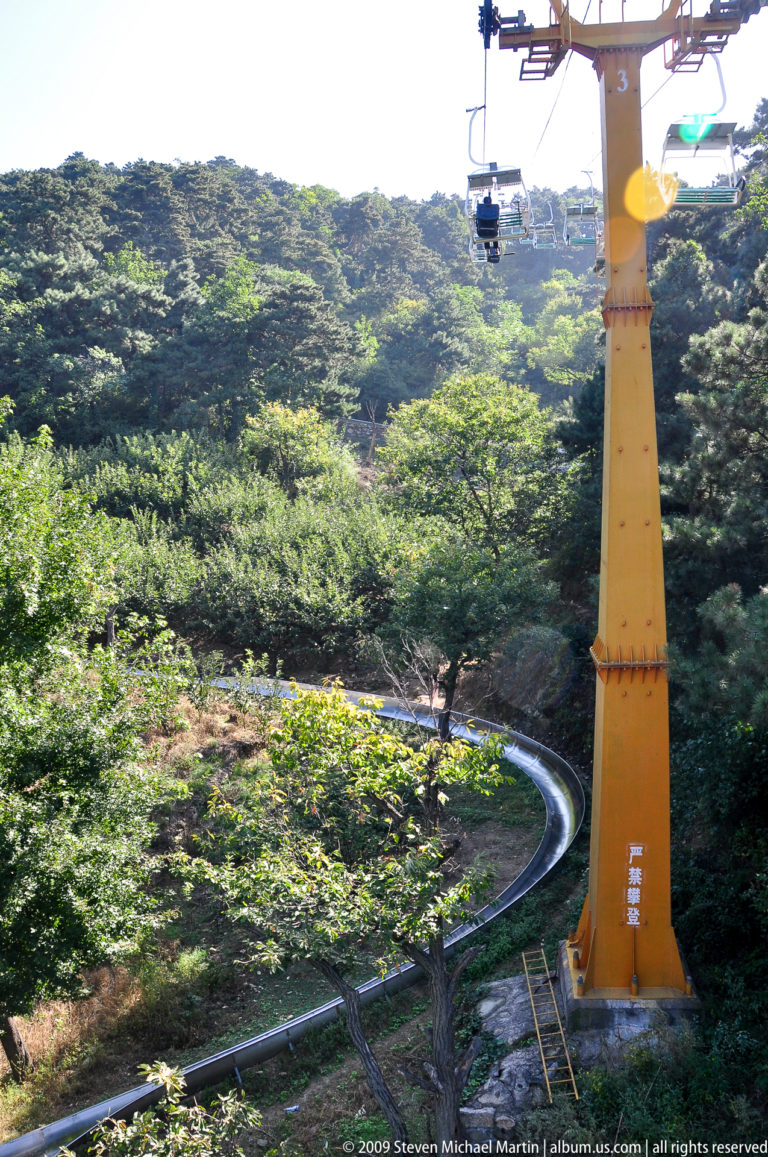 Shiny metal toboggan track curving through dense green forest beside a tall yellow support structure at Mutianyu.