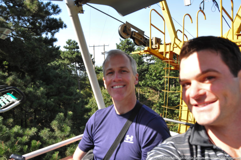 Two male visitors smiling on an open chairlift to the Great Wall at Mutianyu, with lift machinery and pine forest in the background.