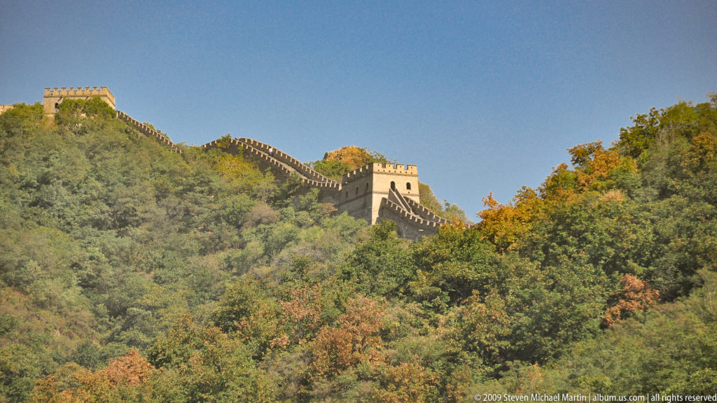 Distant view of a Ming-period Great Wall watchtower and crenellated wall tracing a forested autumn ridge at Mutianyu.