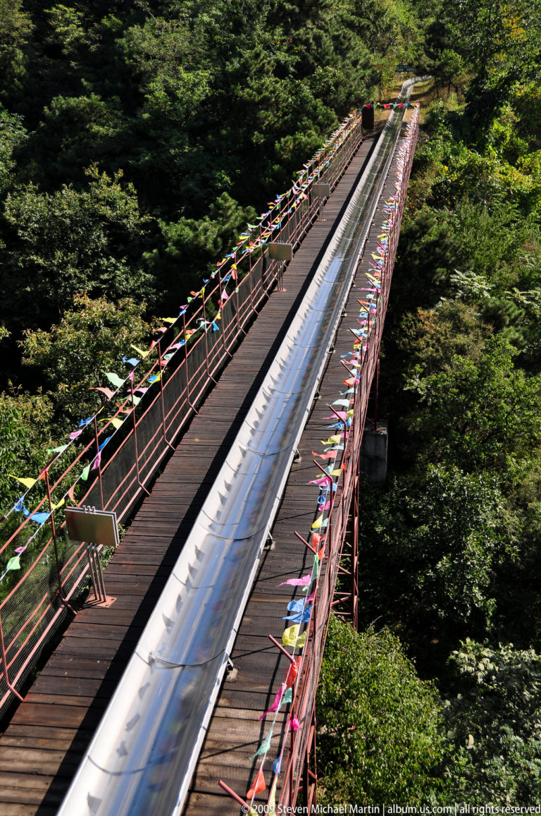 Straight section of stainless-steel alpine slide with adjacent wooden walkway and flags at Mutianyu.