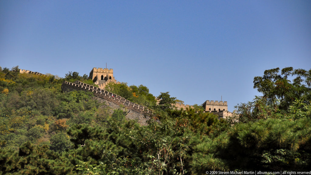 Two Ming‑era Great Wall watchtowers linked by a crenellated brick wall rising along a wooded ridge under a clear blue sky.
