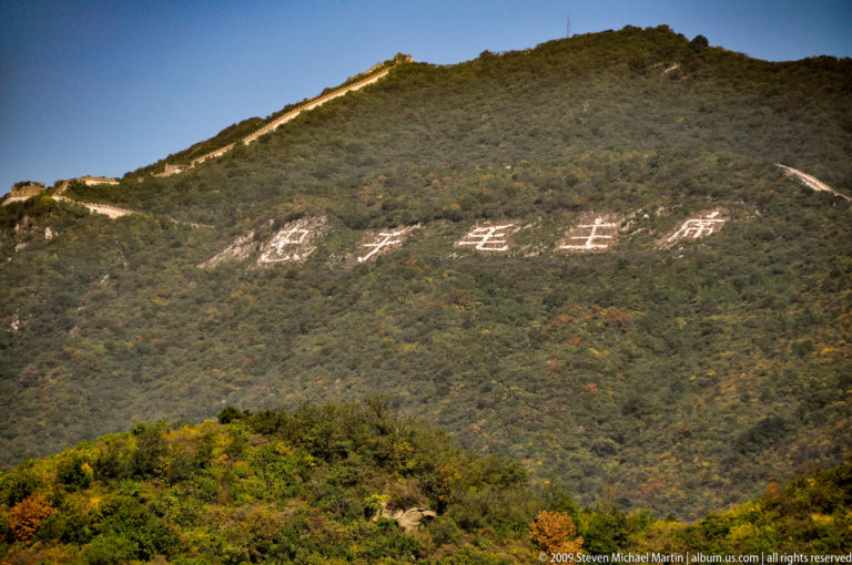 Large white Chinese characters painted on a forested hillside with the Great Wall running along the ridge above at Mutianyu.