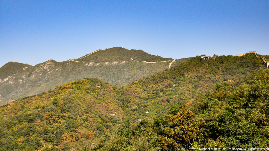 Distant view of the Great Wall running along an autumn mountain ridge above a wooded valley and rocky peaks under a blue sky.