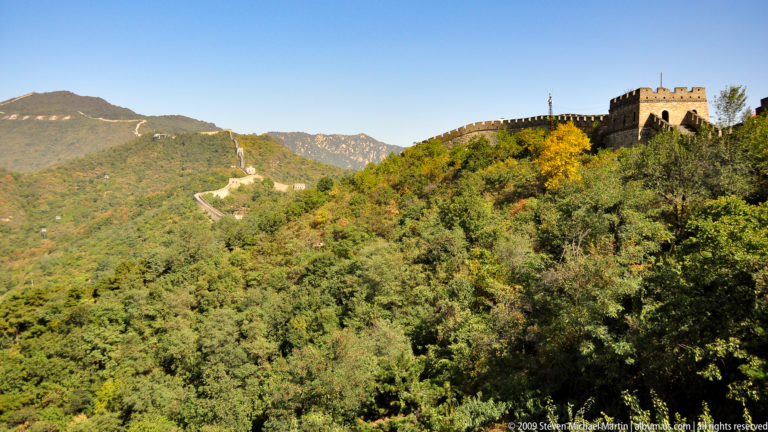 Great Wall watchtower above dense autumn forest, with the wall climbing a steep mountain ridge at Mutianyu.