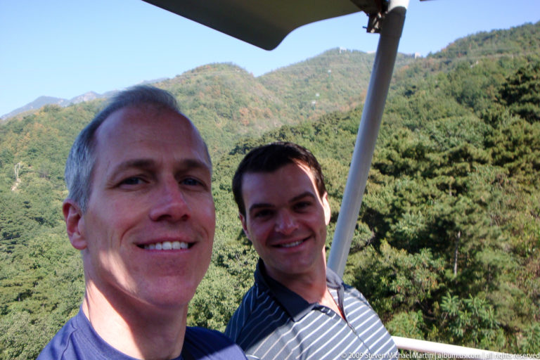Two male visitors smiling on an open chairlift at Mutianyu with forested mountains behind them near the Great Wall.