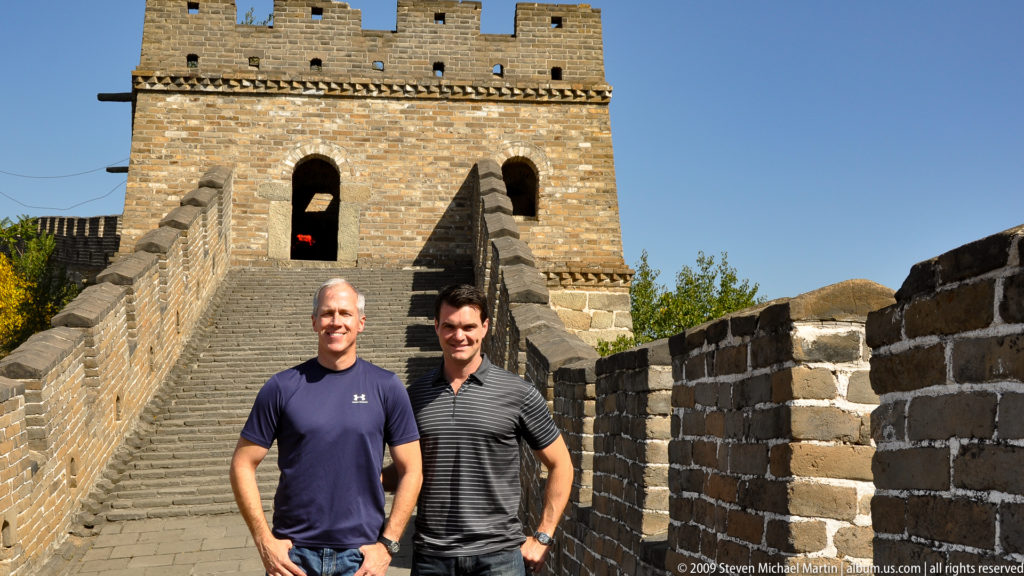 Two visitors stand on wide stone steps in front of a large brick Great Wall watchtower with arched openings and a crenellated roofline at Mutianyu.