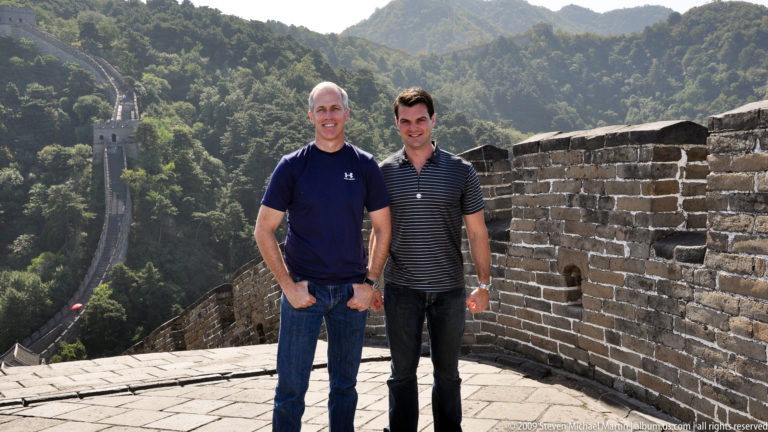 Two visitors stand on a Great Wall platform at Mutianyu beside a brick parapet, with a steep stairway descending into wooded hills behind them.