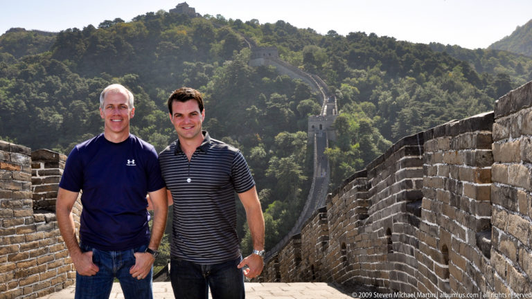 Two visitors stand smiling on a Mutianyu section of the Great Wall of China, with a steep rampart descending to a watchtower in dense forest behind them.
