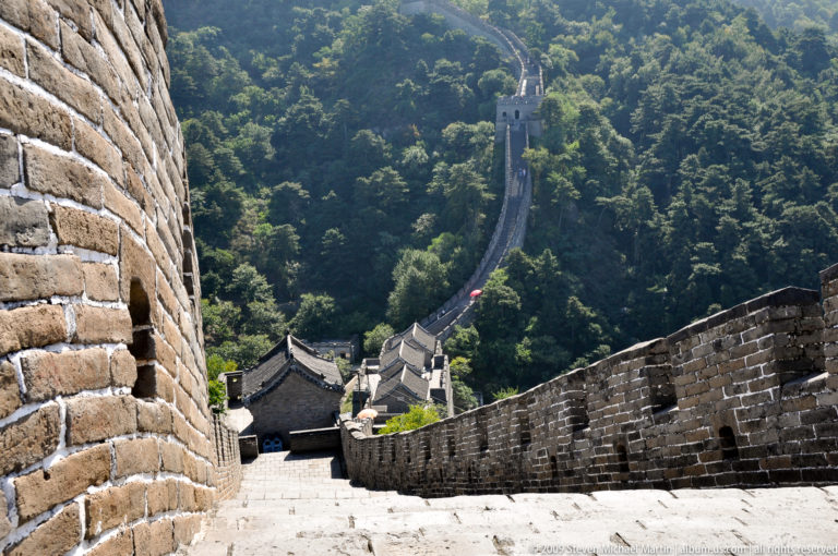 High-angle view down an extremely steep Great Wall stairway between brick parapets, descending toward a watchtower and lower visitor buildings at Mutianyu.