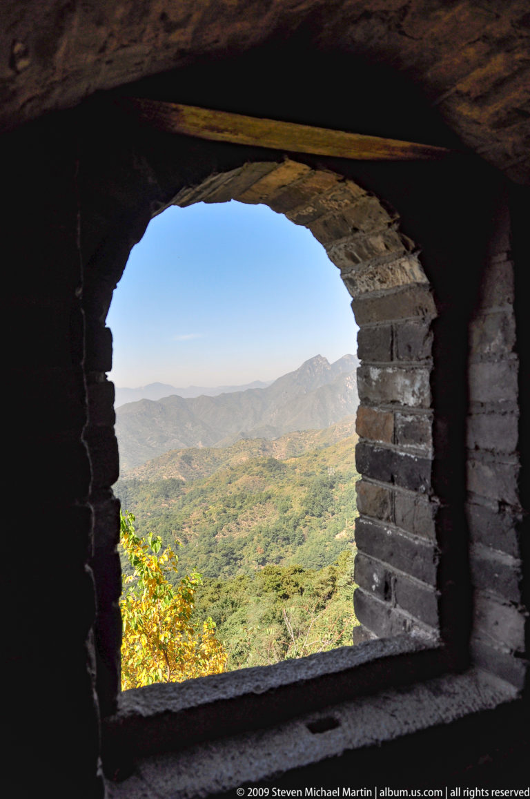 Layered mountain ridges and an autumn valley seen through a dark brick watchtower arch on the Great Wall at Mutianyu.