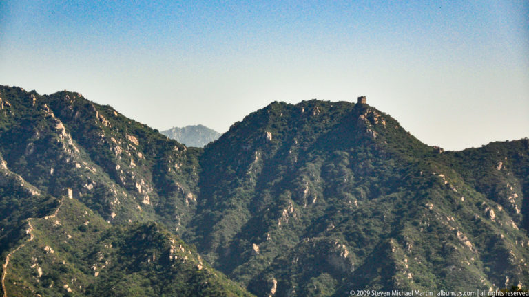 Distant Great Wall watchtower on a high rocky summit above deep mountain valleys.