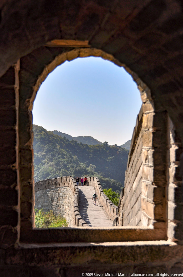 Visitors on a sunlit rampart of the Great Wall at Mutianyu, viewed through the dark brick arch of a Ming watchtower with forested mountains behind.