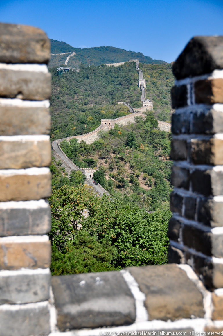 Great Wall of China at Mutianyu curving across a green hillside, with distant watchtowers framed between two out-of-focus brick walls in the foreground.