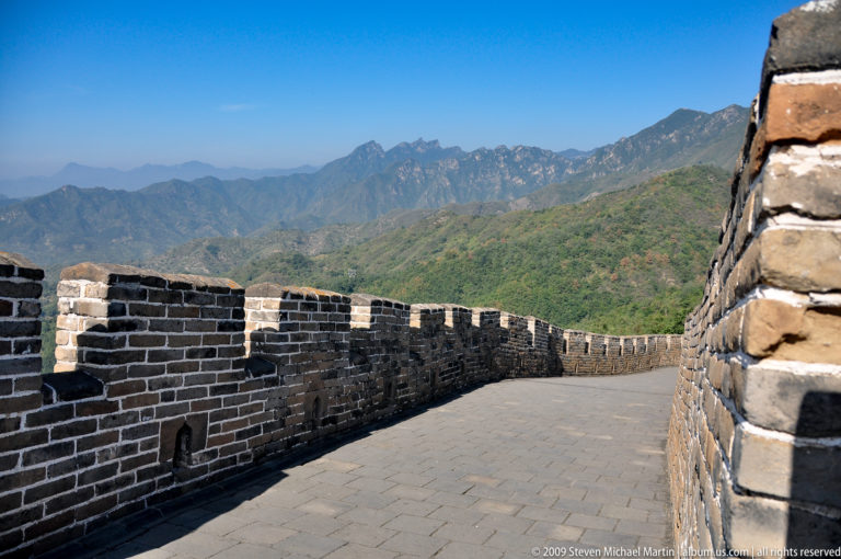 Wide brick-paved Great Wall walkway at Mutianyu curving between crenellated parapets toward distant mountains under a clear blue sky.