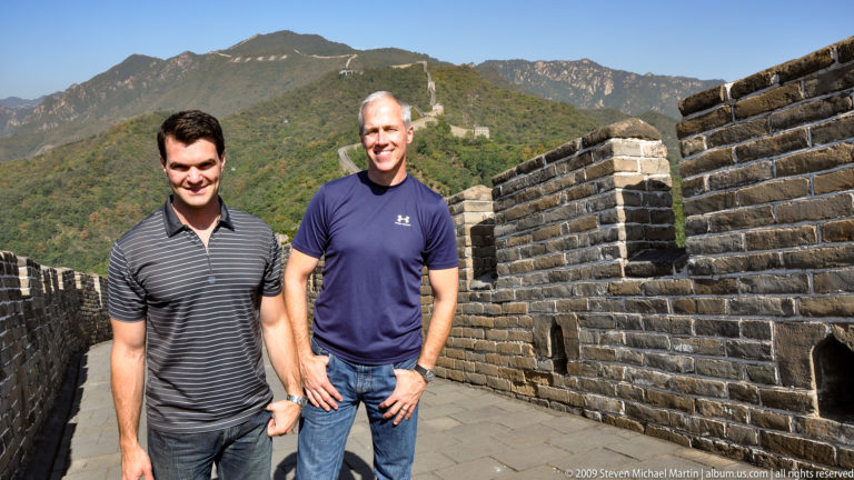 Two male visitors stand smiling on the brick-paved Great Wall rampart at Mutianyu, with crenellated parapets and a long wall with watchtowers stretching over a wooded mountain ridge behind them.