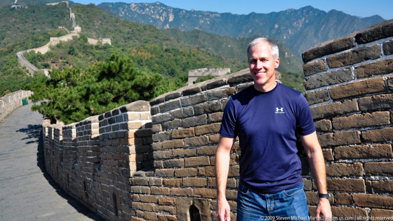 Man in a blue shirt standing beside a brick parapet on the Great Wall at Mutianyu, with watchtowers and a mountain ridge behind him.