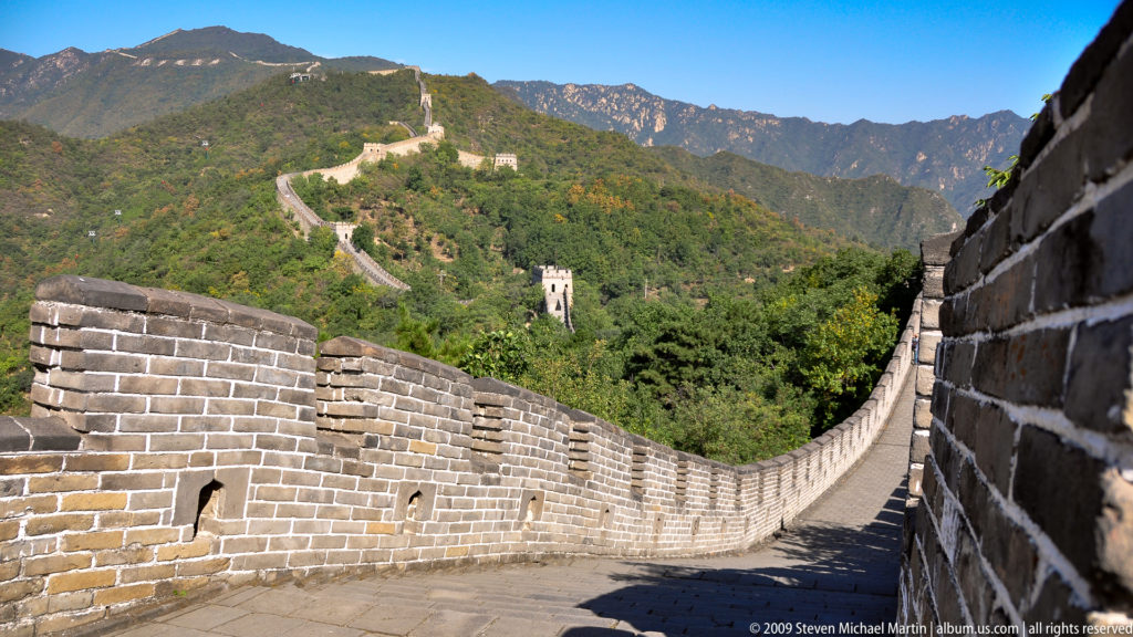 Wide view of the Great Wall at Mutianyu, its brick-paved rampart dipping sharply through a mountain saddle and rising to watchtowers on the far ridge.