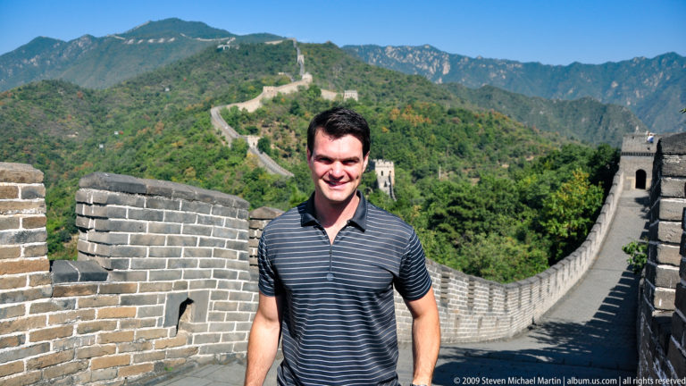 Man standing and smiling beside a parapet on the Great Wall at Mutianyu, with the brick-paved rampart winding over the surrounding mountain ridges toward multiple watchtowers.