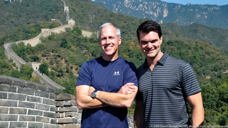 Two men stand on a restored section of the Great Wall at Mutianyu, with the brick-paved rampart and distant watchtowers winding along forested mountain ridges behind them.