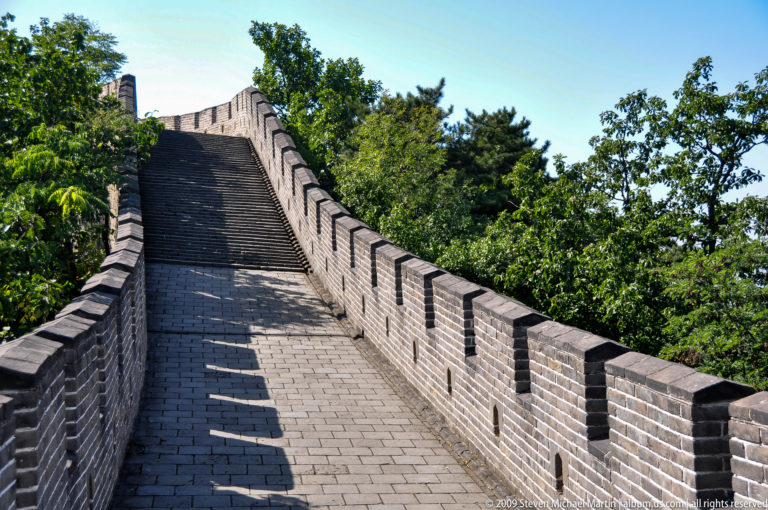 Steep, empty brick-paved Great Wall rampart with restored crenellated parapets ascending through dense trees at Mutianyu.