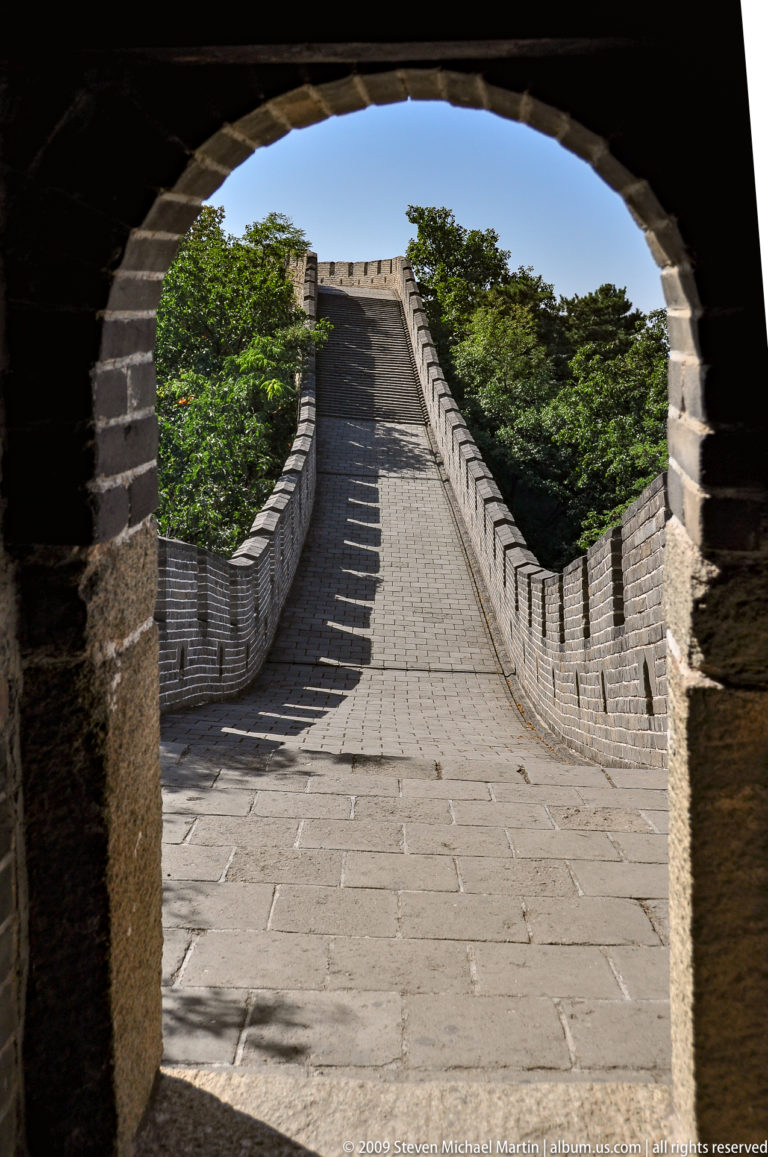View from inside a Great Wall watchtower at Mutianyu, looking out through a dark arched opening onto a steep brick-paved rampart lined with crenellated parapets.