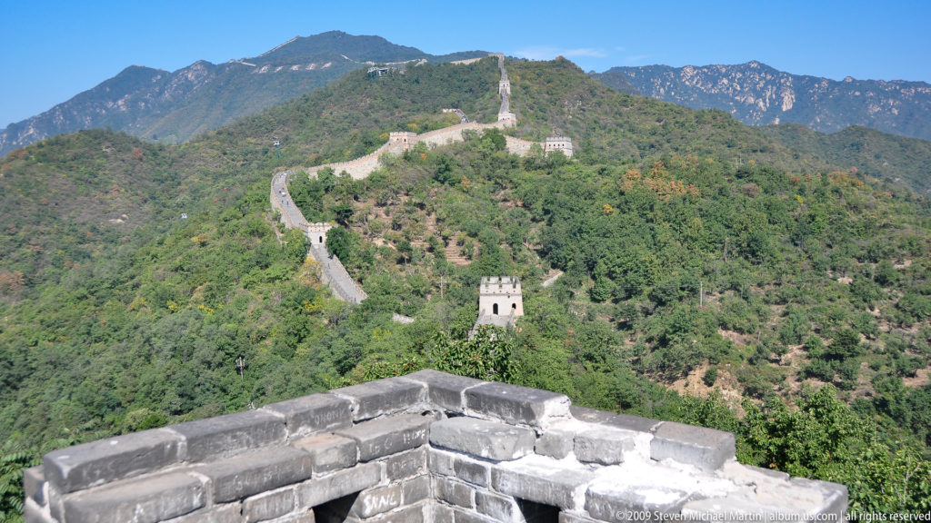 View from a crenellated Mutianyu battlement looking along the Great Wall and its watchtowers as they follow green mountain ridges.