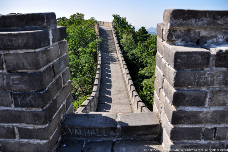 Steep, empty Great Wall rampart at Mutianyu rising through trees, viewed between close brick crenellations.