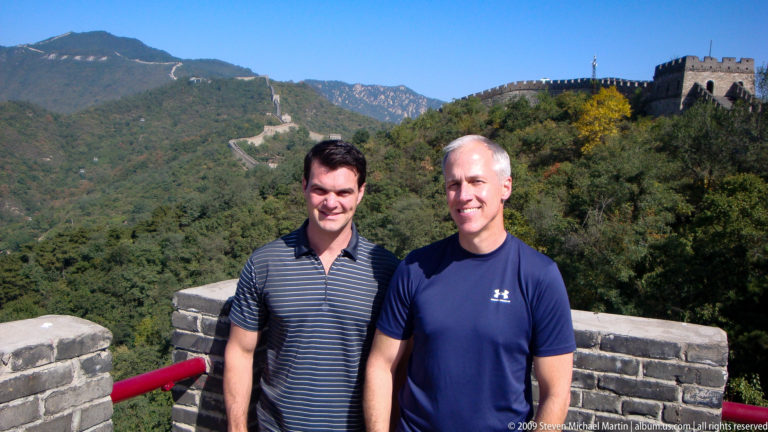 Two male visitors stand beside a brick parapet on the Great Wall at Mutianyu with watchtowers stepping along a steep, forested mountain ridge in the background.