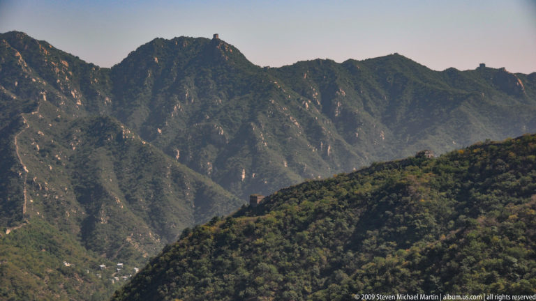 Distant mountain ridges with a small Great Wall watchtower on a wooded crest, fading into hazy background peaks.