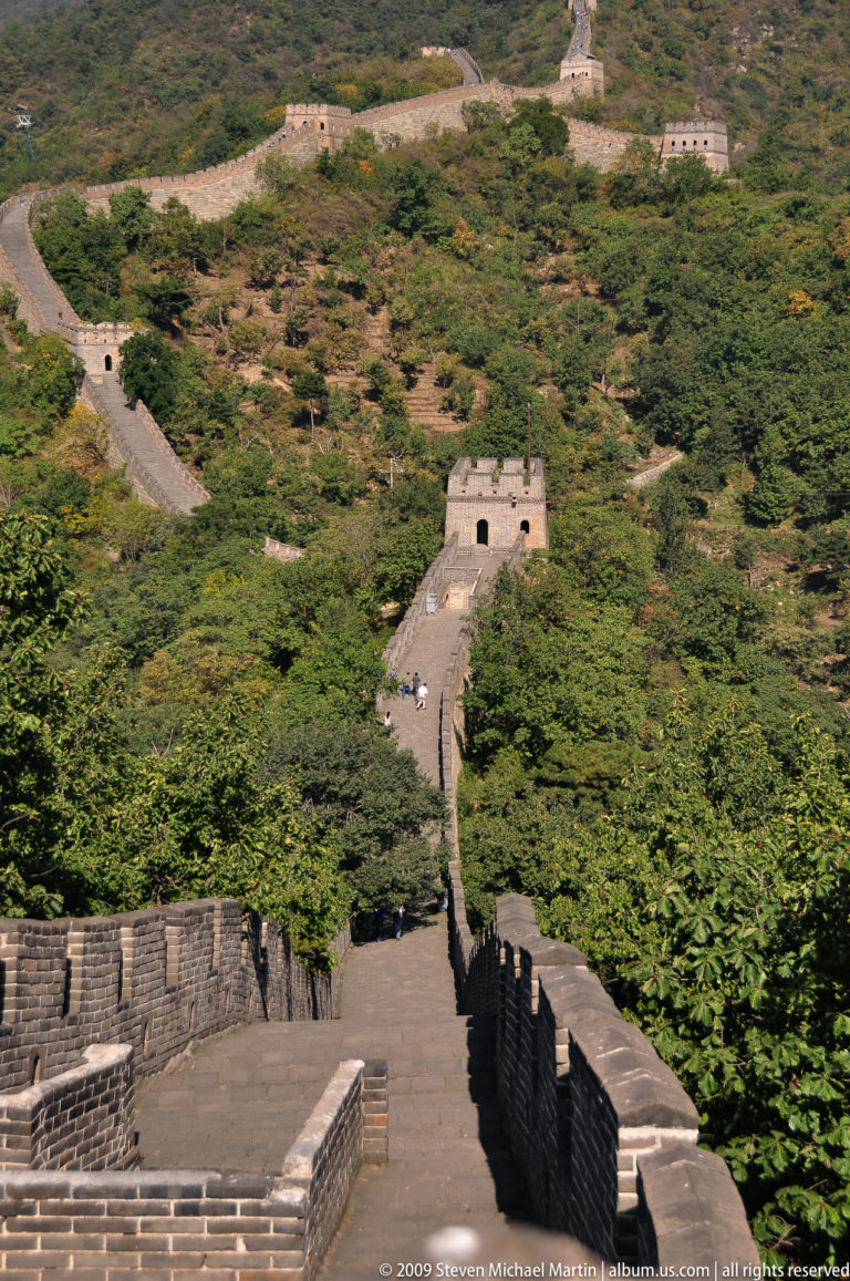 Ming dynasty Great Wall watchtower at Mutianyu with a narrow, steep rampart running through dense forest on a mountain slope.