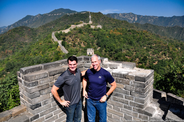 Two men stand on a low-walled platform at Mutianyu with the Great Wall and several watchtowers winding along the distant mountain ridge.