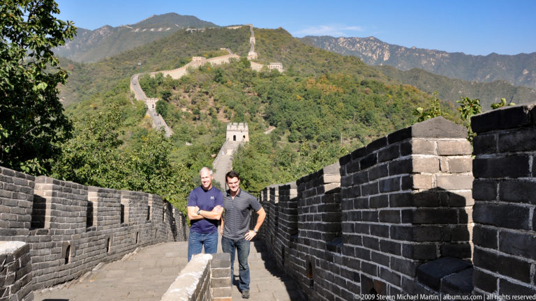 Two visitors standing on a descending brick walkway of the Great Wall at Mutianyu, enclosed by high crenellated parapets with a watchtower and wall stretching over the mountain in the distance.