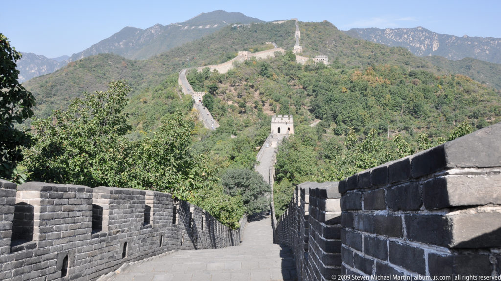 Wide view of an empty brick-paved section of the Great Wall at Mutianyu descending between crenellated parapets toward a central watchtower, with the wall continuing up a distant mountain ridge.