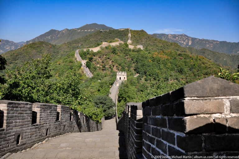 Panoramic view of the Mutianyu Great Wall descending from the foreground and crossing distant wooded ridges, with evenly spaced watchtowers along the high mountain route.
