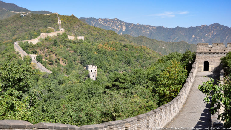 Curving Great Wall rampart at Mutianyu descending into a forested valley with watchtowers on the surrounding ridges.