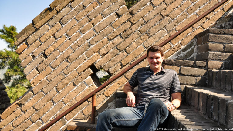 Male visitor resting on steep brick stairs beside a metal handrail on the Great Wall at Mutianyu.