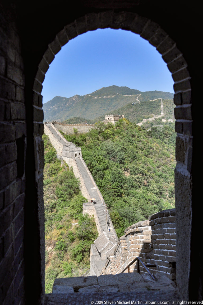 View through a dark watchtower doorway at Mutianyu, framing a sunlit stretch of the Great Wall with crenellated ramparts and several towers crossing a distant mountain slope.