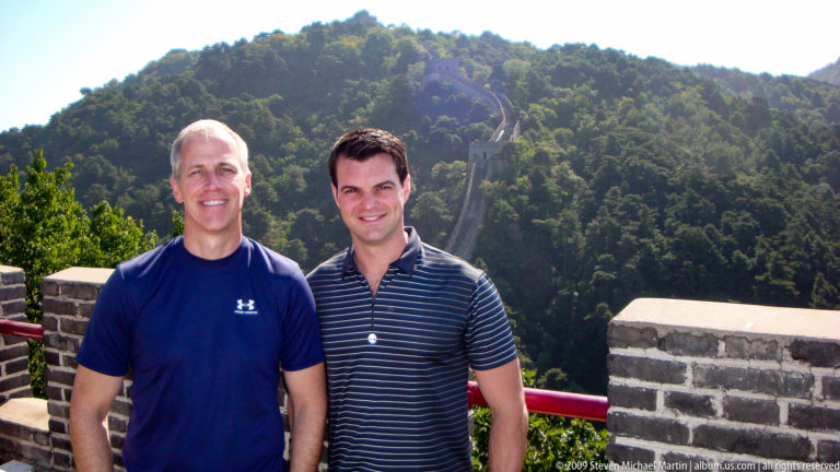 Two male visitors standing and smiling at a parapet on the Mutianyu Great Wall, with a steep restored Ming brick rampart and watchtower rising along the wooded mountain slope behind them.