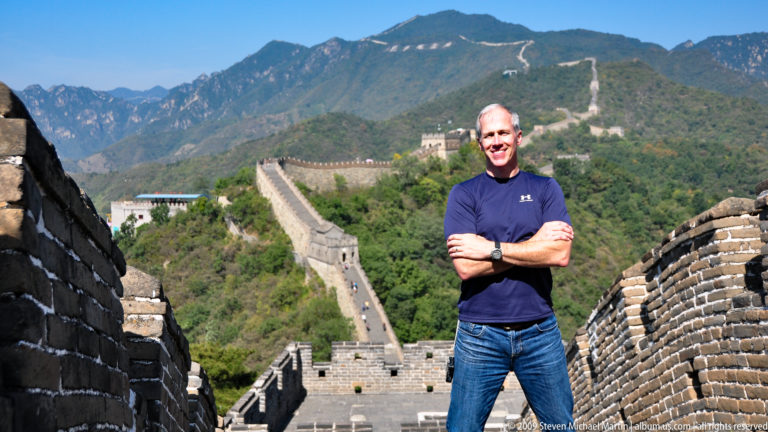 Man in blue standing with arms crossed on the Great Wall at Mutianyu, with restored Ming-era ramparts and watchtowers stretching over the mountains behind him.