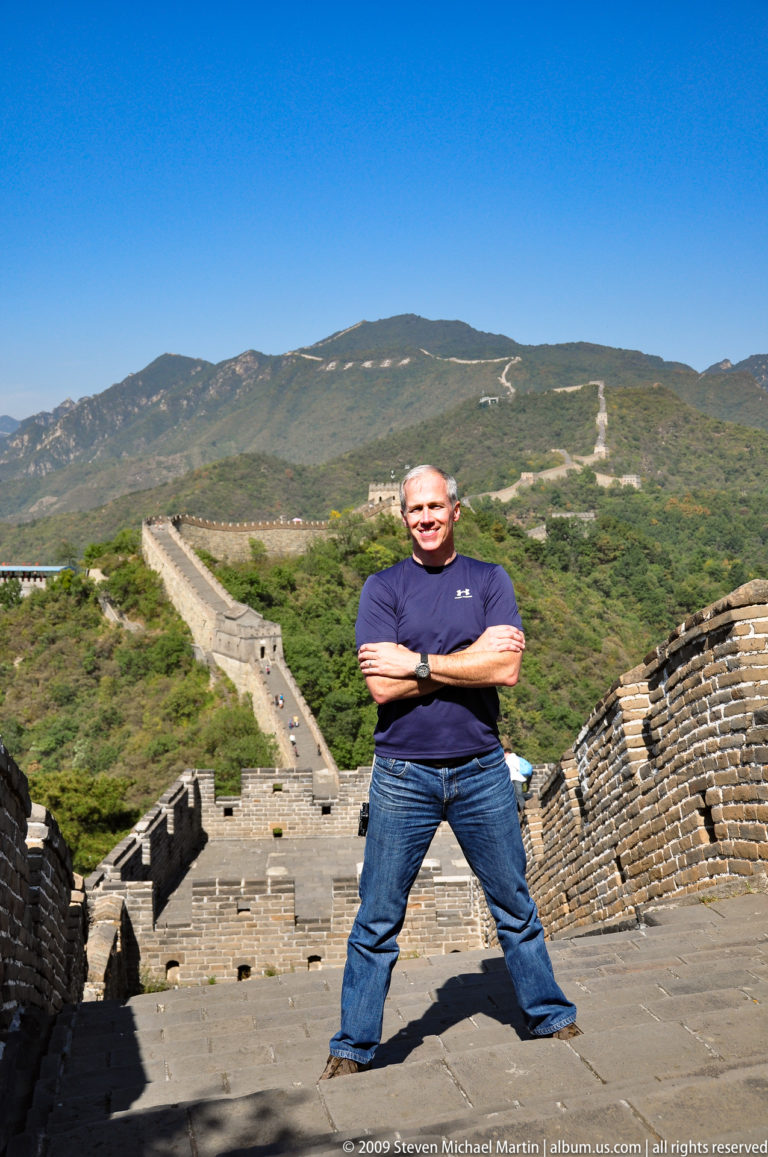 Man with arms crossed standing on the Mutianyu Great Wall, with crenellated ramparts and watchtowers running along a mountain ridge behind him.