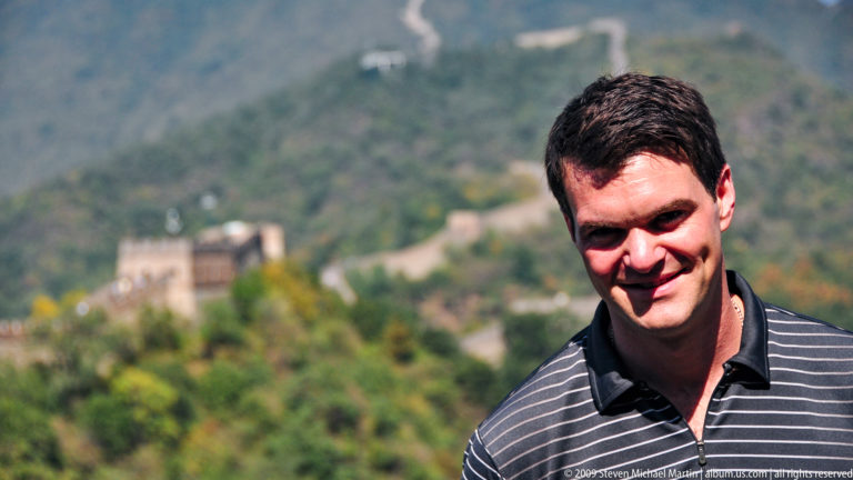 Smiling male visitor in close-up with a blurred Great Wall watchtower and forested slope in the background at Mutianyu.