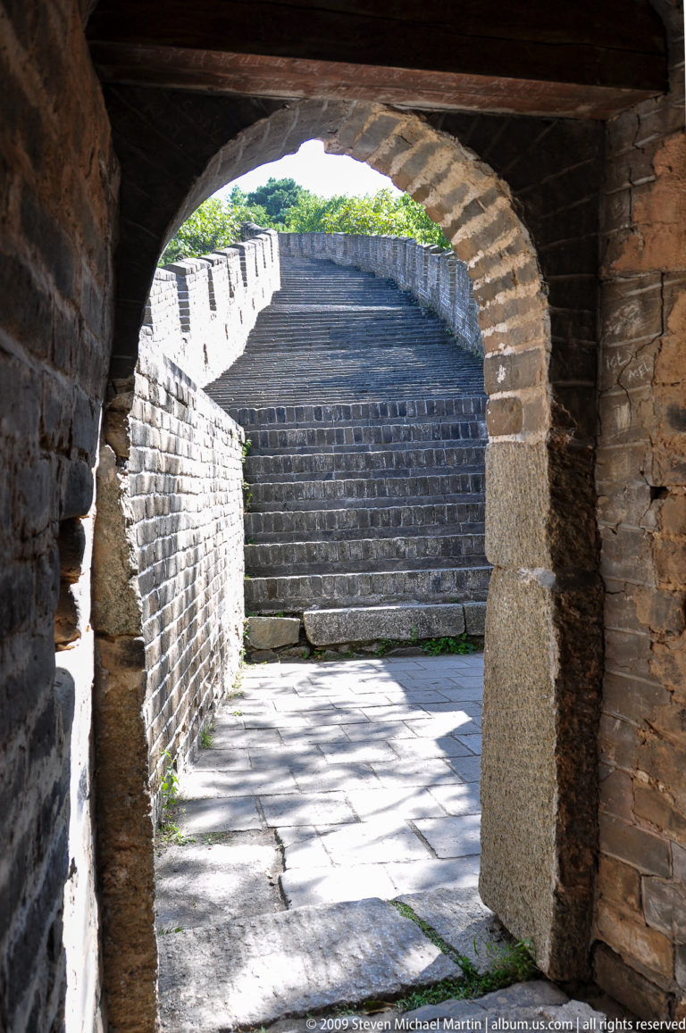 Sunlit interior of a Mutianyu Great Wall watchtower, showing a steep stone stair rising beside a thick brick arched doorway.