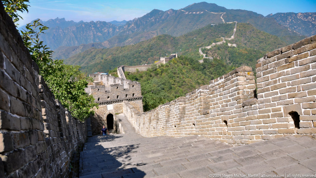 View down a wide brick rampart of the Great Wall at Mutianyu toward a square watchtower, with crenellated parapets and additional towers visible along the mountain ridge behind.