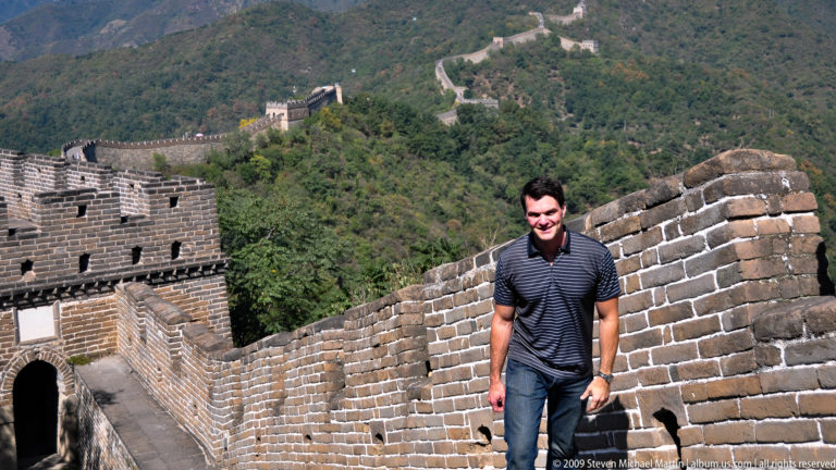 Male visitor in a striped shirt climbing a steep brick rampart of the Mutianyu Great Wall with distant watchtowers along the mountain ridge behind him.