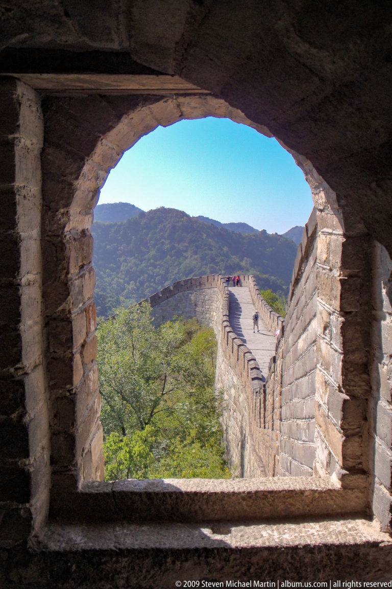 View through a dark brick watchtower arch at Mutianyu, showing the Great Wall’s narrow, crenellated rampart curving toward a distant tower with small visitors on the walkway.