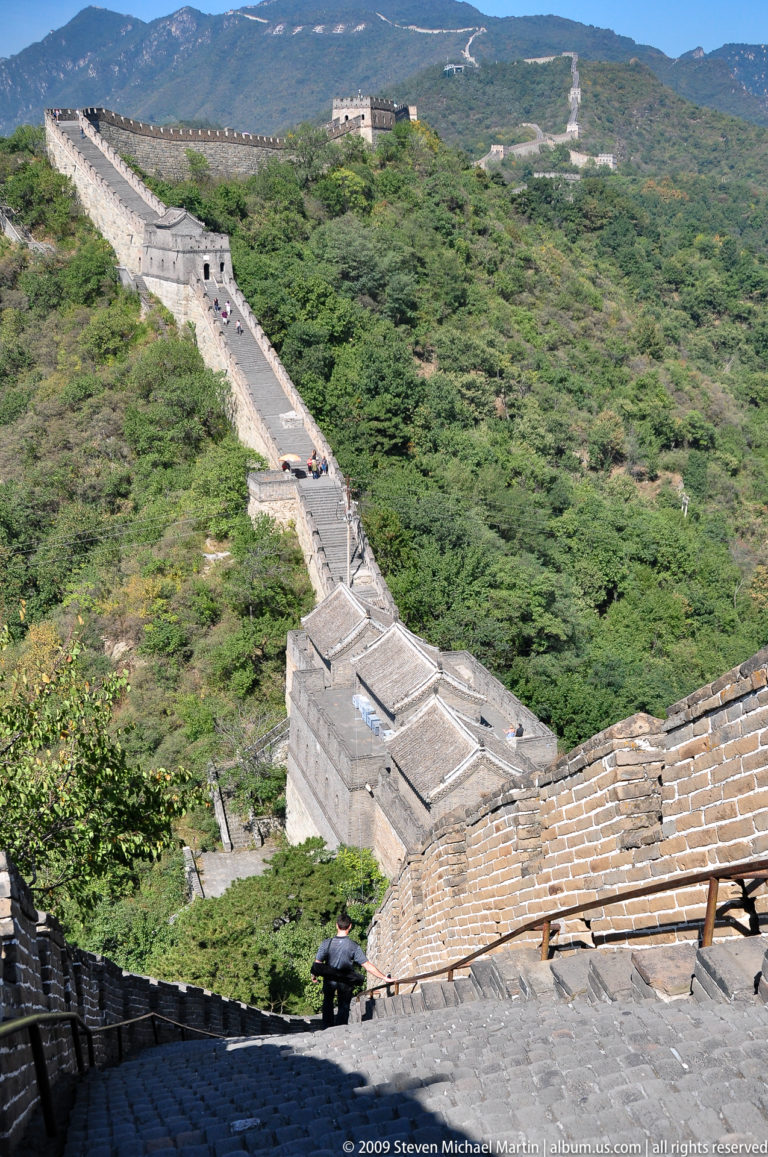 High-angle view of gray-tiled Great Wall watchtower roofs stepping down a slope beside wall stairs, surrounded by dense forest at Mutianyu.