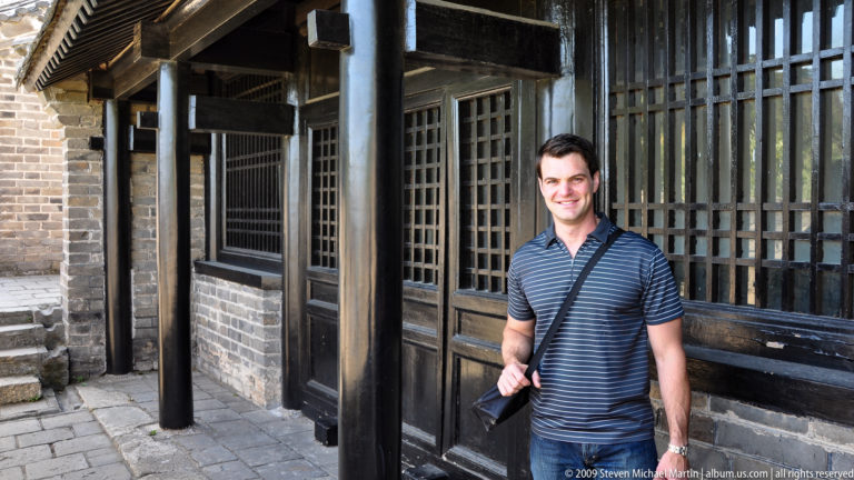 Male visitor with a camera standing beside a timber-fronted building with lattice windows and a gray-brick base near the Great Wall at Mutianyu.